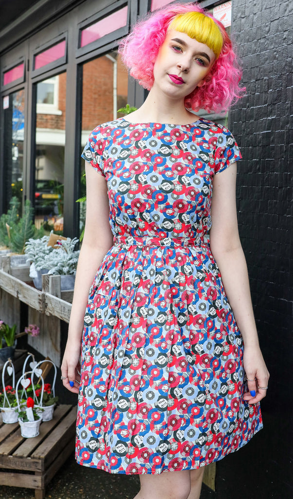 Lottie is stood in front of a flower shop wearing the vinyl record print slate grey tea party dress, they have pink and yellow curly hair. They are facing forward posing with with both hands resting by her side with her head tilted to one side. Photo is cropped from the knees up.