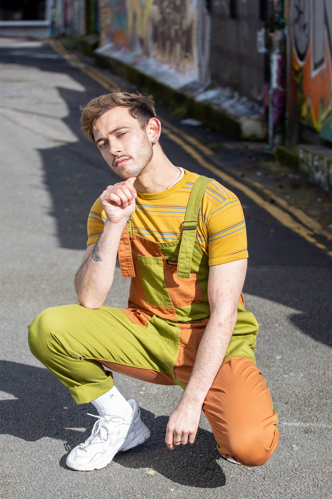 Zach is crouching down in a graffitied alleyway wearing the sunset stripe retro tshirt with green and orange dungarees and white trainers. He is facing the camera posing with one hand under his chin.